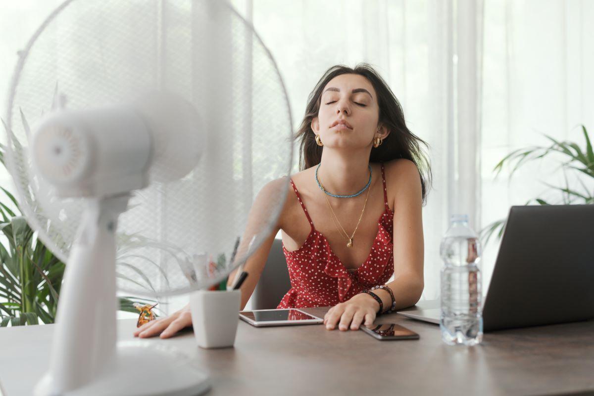 Woman sitting at a table with a fan blowing on her face, illustrating the importance of home inspections in the hot Dallas/Fort Worth summer season to ensure HVAC efficiency and overall home comfort