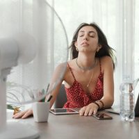 Woman sitting at a table with a fan blowing on her face, illustrating the importance of home inspections in the hot Dallas/Fort Worth summer season to ensure HVAC efficiency and overall home comfort